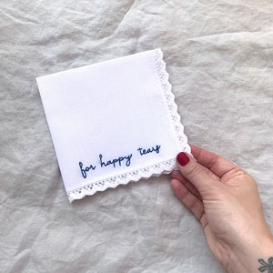Image shows a white handkerchief with a detailed edge, folded into quarters. The text reads for happy tears in blue embroidery thread. A hand is holding onto the bottom right corner of the hanky.