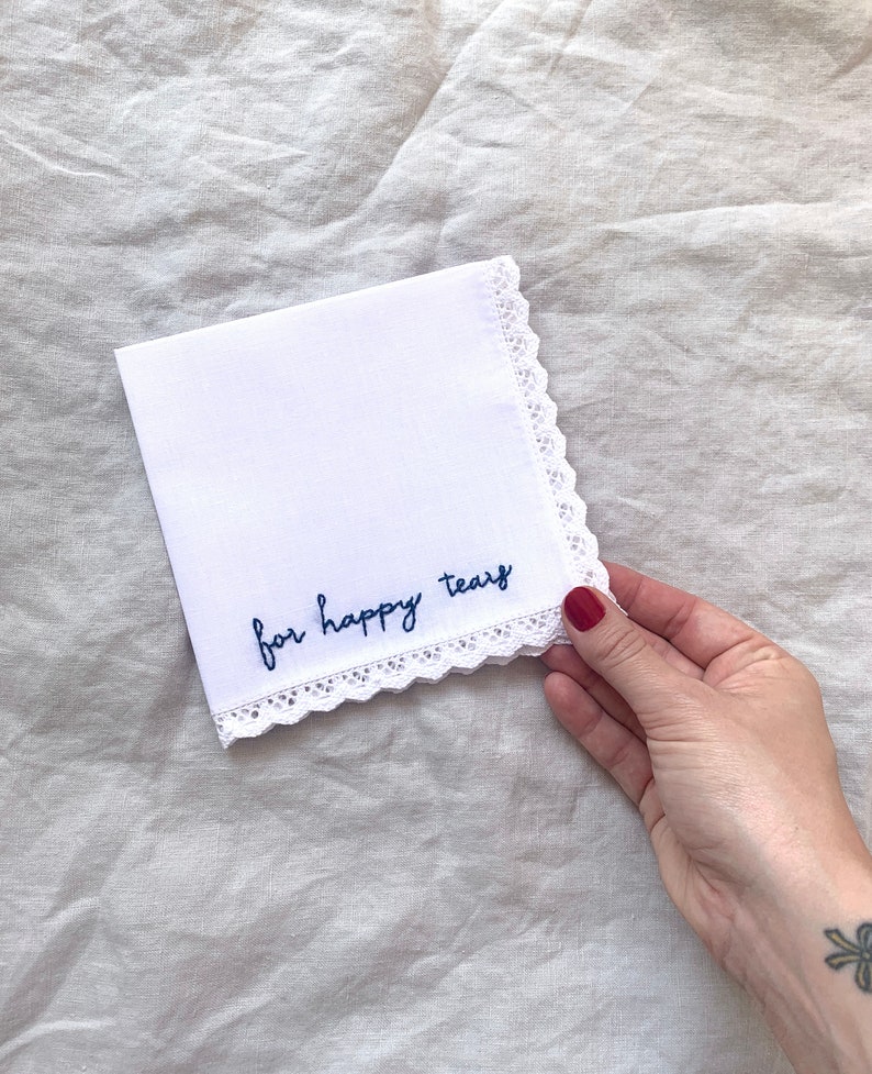 Image shows a white handkerchief with a detailed edge, folded into quarters. The text reads for happy tears in blue embroidery thread. A hand is holding onto the bottom right corner of the hanky.