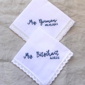 Image shows two white handkerchiefs with detailed edges, folded into quarters and overlapping one another. The text shows "Mrs. Gorman" and "Mrs. Betschart" along with a date on each, embroidered in blue thread.