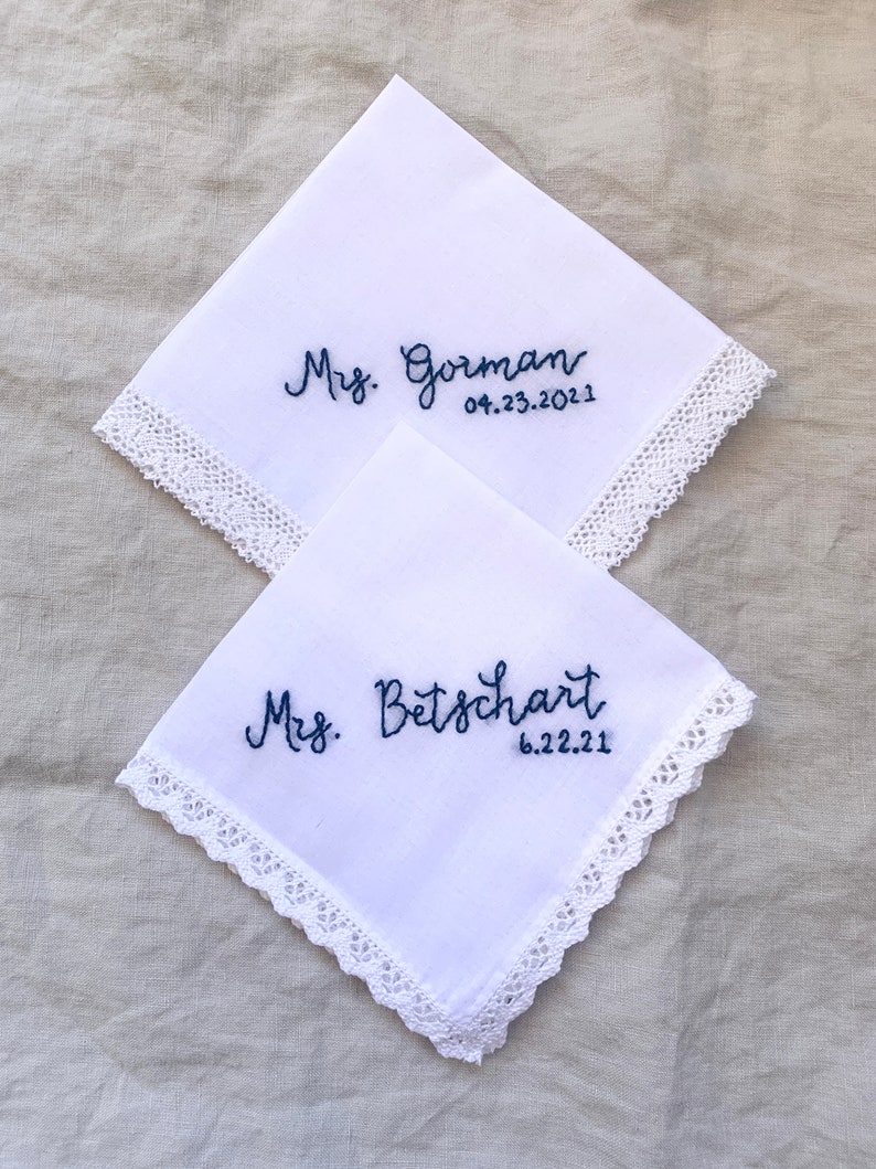 Image shows two white handkerchiefs with detailed edges, folded into quarters and overlapping one another. The text shows "Mrs. Gorman" and "Mrs. Betschart" along with a date on each, embroidered in blue thread.