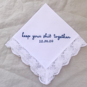 Image shows a white handkerchief with a detailed edge, folded into quarters. The text reads keep your shit together in blue embroidery thread, with the date underneath.