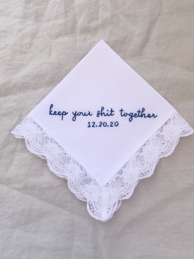 Image shows a white handkerchief with a detailed edge, folded into quarters. The text reads keep your shit together in blue embroidery thread, with the date underneath.