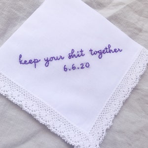 Image shows a white handkerchief with a detailed edge, folded into quarters. The text reads keep your shit together in purple embroidery thread, with the date underneath.