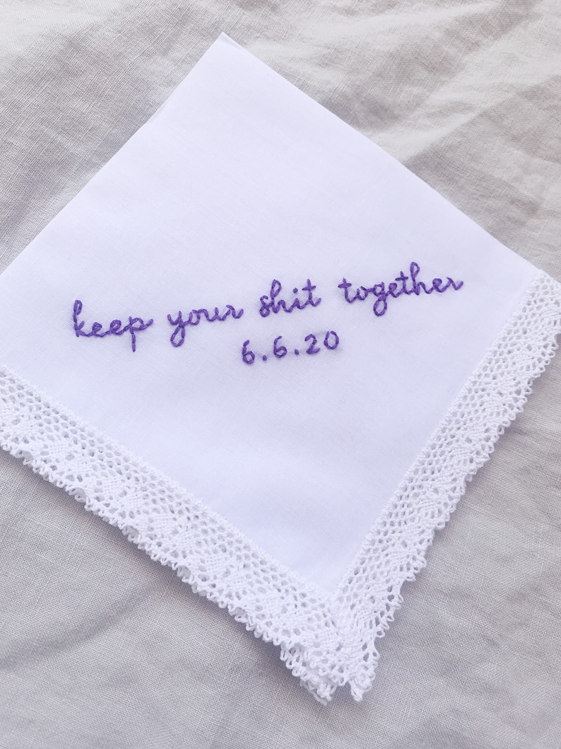 Image shows a white handkerchief with a detailed edge, folded into quarters. The text reads keep your shit together in purple embroidery thread, with the date underneath.