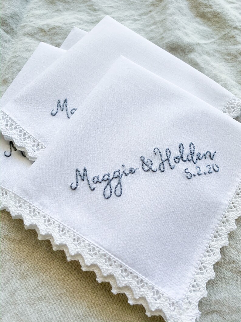 Image shows a stack of white handkerchiefs with a detailed edge, folded into quarters. The text shows "Maggie & Holden" along with the date, embroidered in gray thread.