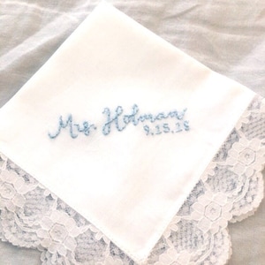 Image shows a white handkerchief with a detailed edge, folded into quarters. The text shows "Mrs. Holman" along with the date, embroidered in light blue thread.