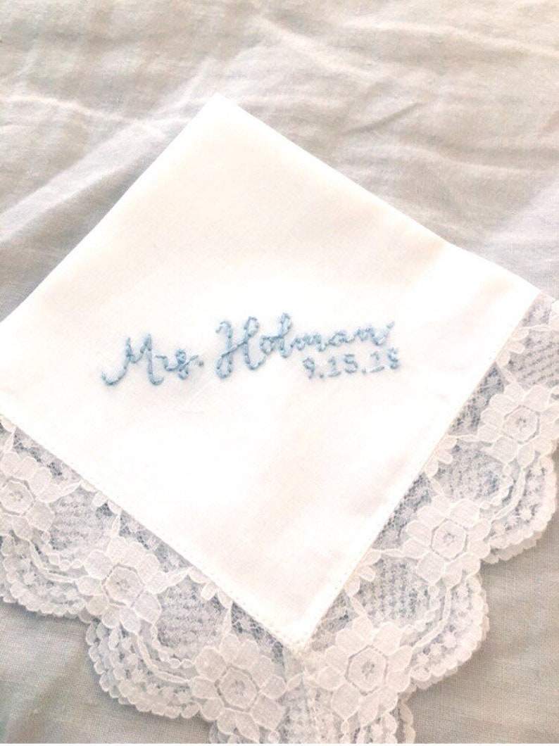 Image shows a white handkerchief with a detailed edge, folded into quarters. The text shows "Mrs. Holman" along with the date, embroidered in light blue thread.