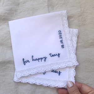 Image shows two layered white handkerchiefs with a detailed edge, folded into quarters. The text reads for happy tears in blue embroidery thread, and the date is embroidered along the perpendicular edge.