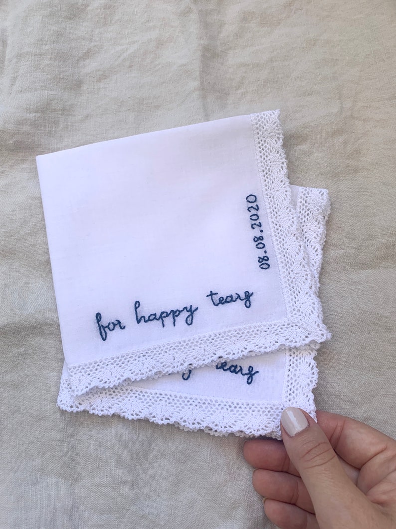 Image shows two layered white handkerchiefs with a detailed edge, folded into quarters. The text reads for happy tears in blue embroidery thread, and the date is embroidered along the perpendicular edge.