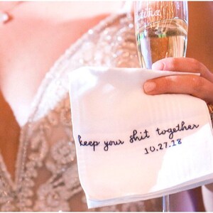 Image shows a bride holding a white handkerchief with line deatil in the fabric, as well as a glass of champagne. The text reads keep your shit together in blue embroidery thread, with the date underneath.