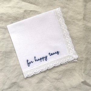 Image shows a white handkerchief with a detailed edge, folded into quarters. The text reads for happy tears in blue embroidery thread.