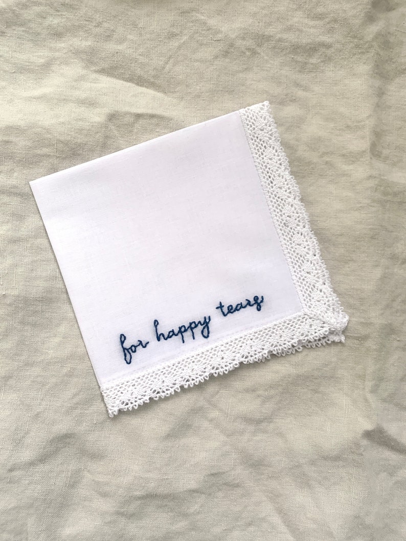 Image shows a white handkerchief with a detailed edge, folded into quarters. The text reads for happy tears in blue embroidery thread.
