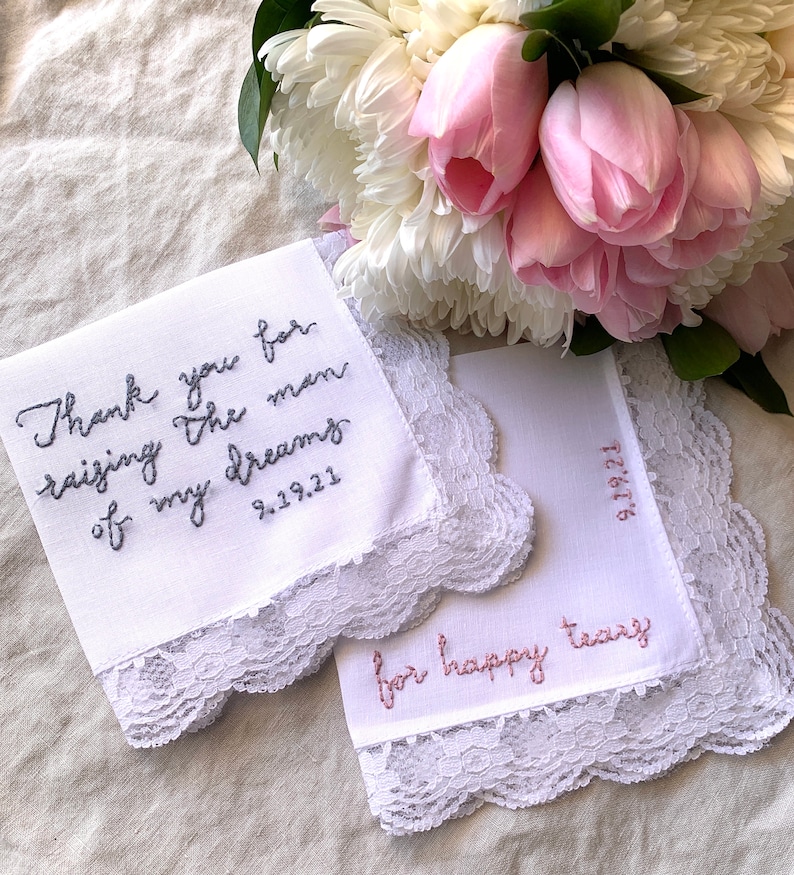 Image shows a white handkerchief with a detailed edge, folded into quarters. The text is the standard message for this listing, with a date added on, in greay thread. Another hankie from a different listing is also in the photo, along with flowers.