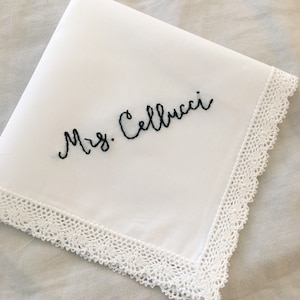 Image shows a white handkerchief with a detailed edge, folded into quarters. The text shows "Mrs. Cellucci", embroidered in black thread.