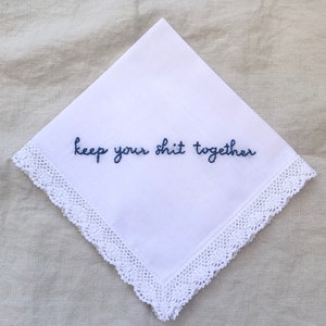 Image shows a white handkerchief with a detailed edge, folded into quarters. The text reads keep your shit together in blue embroidery thread.