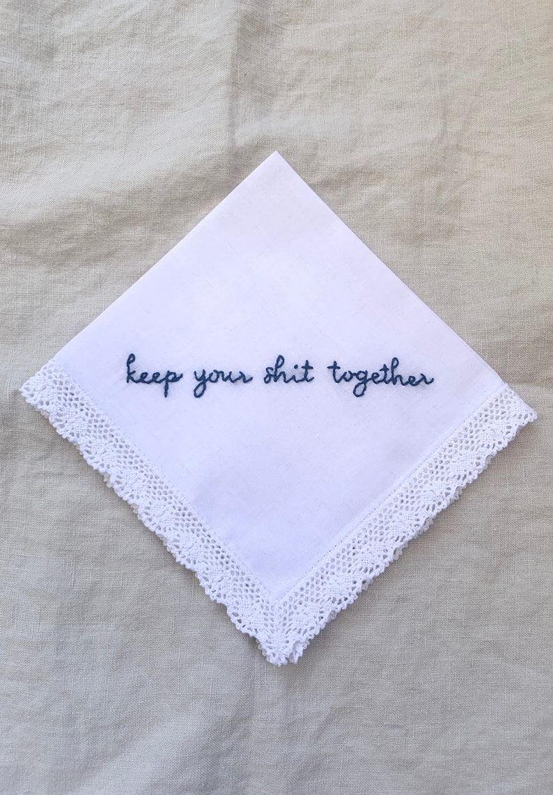 Image shows a white handkerchief with a detailed edge, folded into quarters. The text reads keep your shit together in blue embroidery thread.
