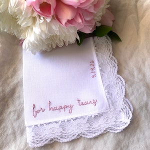 Image shows a white handkerchief with a detailed edge, folded into quarters. The text reads for happy tears in pink embroidery thread, and the date is embroidered along the perpendicular edge. A bouquet of flowers rests at the top of the hanky.