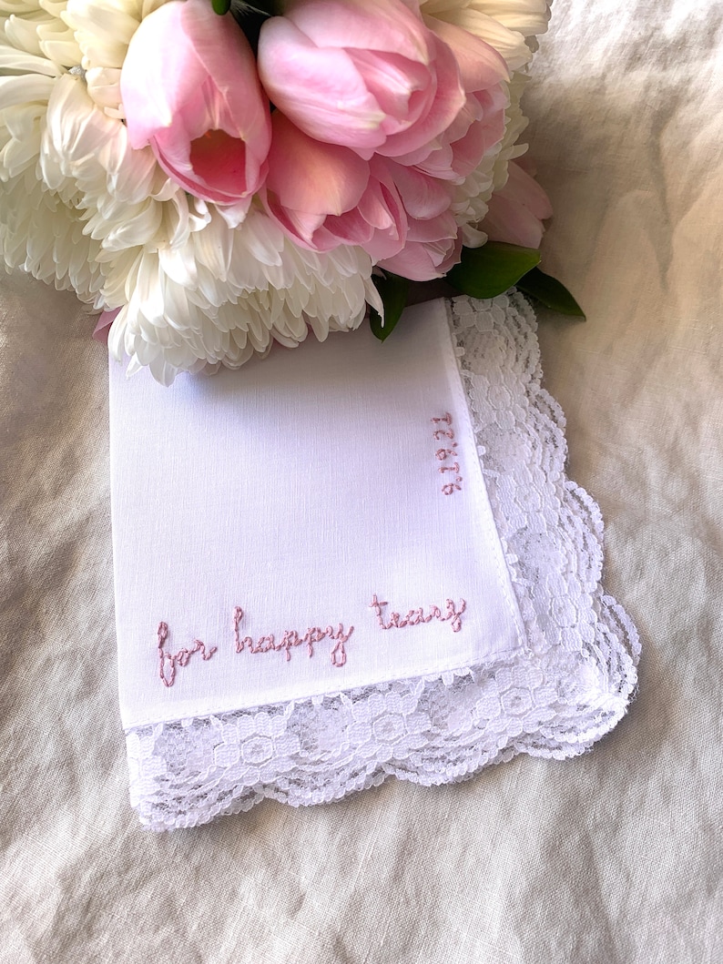 Image shows a white handkerchief with a detailed edge, folded into quarters. The text reads for happy tears in pink embroidery thread, and the date is embroidered along the perpendicular edge. A bouquet of flowers rests at the top of the hanky.