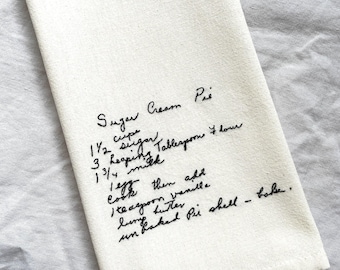 May include: A white cotton kitchen towel featuring a handwritten recipe for Sugar Cream Pie in black ink. The text lists ingredients including 1 1/2 cups sugar, 3 heaping tablespoons flour, 1 3/4 cups milk, 1 egg, 1 teaspoon vanilla, and butter.