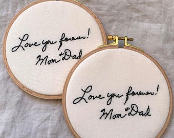 May include: Two circular wooden embroidery hoops containing off-white fabric. Each hoop features black hand-stitched cursive text that reads Love you forever! Mom + Dad. The hoops have gold-toned metal tightening hardware at the top.