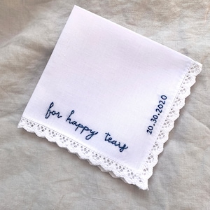 Image shows a white handkerchief with a detailed edge, folded into quarters. The text reads for happy tears in blue embroidery thread. The date is embroidered along the perpendicular edge.