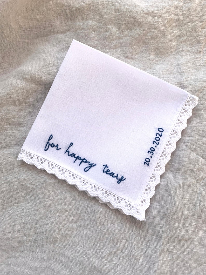 Image shows a white handkerchief with a detailed edge, folded into quarters. The text reads for happy tears in blue embroidery thread. The date is embroidered along the perpendicular edge.