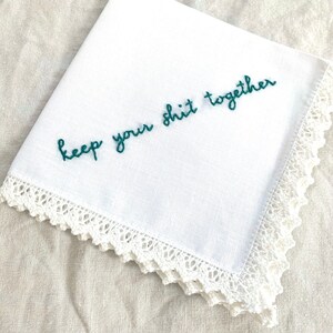 Image shows a white handkerchief with a detailed edge, folded into quarters. The text reads keep your shit together in green embroidery thread.