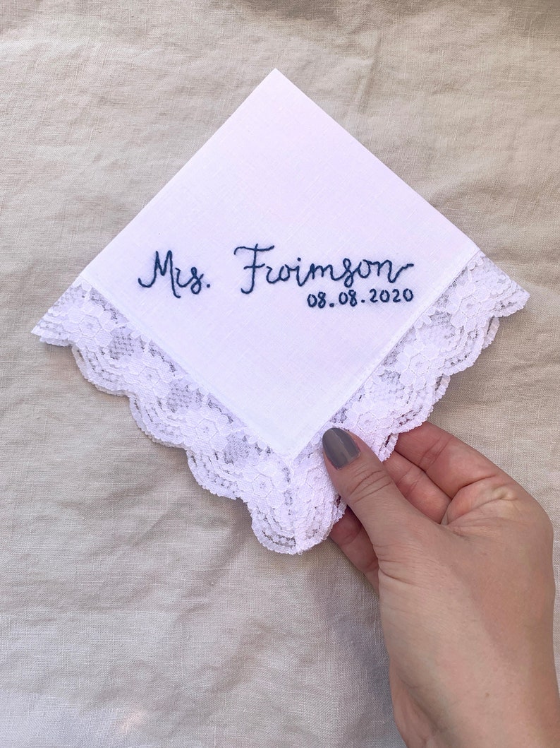 Image shows a white handkerchief with a detailed edge, folded into quarters. The text shows "Mrs. Froimson" along with the date, embroidered in blue thread.