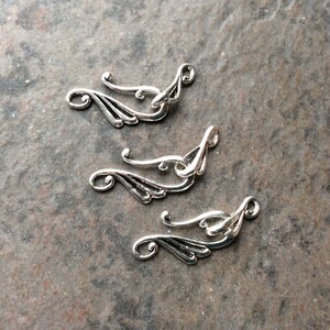 May include: Close-up of four silver-colored metal jewelry findings. Each finding has a curved, wing-like design with a loop at one end and a decorative swirl. The findings are arranged on a textured, gray surface.