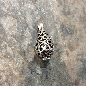 May include: A silver filigree pendant in the shape of a teardrop. The pendant has a hinged opening and is designed to hold small items, such as essential oils or crystals.