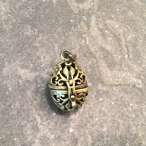 May include: A gold-toned metal pendant shaped like an egg with an intricate, openwork design. The pendant has a hinged lid and is designed to hold small items.