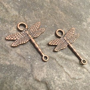 May include: Two copper-colored dragonfly charms with a loop at the top and a jump ring at the bottom. The dragonflies have detailed wings and a textured body.