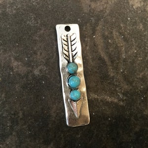 May include: Silver arrow charm with three turquoise stones. The arrow is on a silver rectangle with a textured surface.