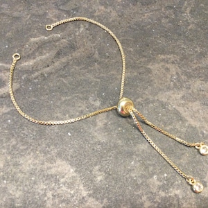 May include: Gold-toned adjustable bracelet chain with a box chain design. The bracelet features a sliding bead for adjustment and small, clear, round charms at the ends of the dangling chains. The bracelet is displayed on a gray surface.