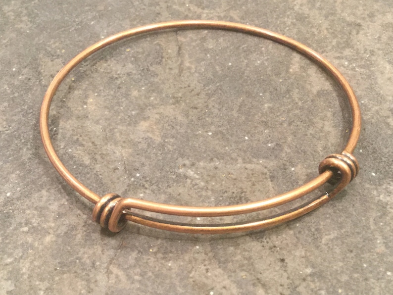 May include: A copper expandable bangle bracelet with a simple design. The bracelet is made of a single piece of wire that is bent into a circle and then twisted at the ends to create a secure closure.
