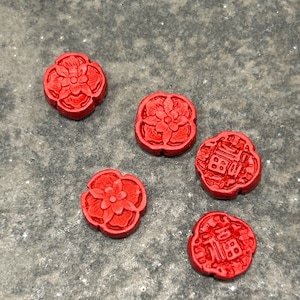 Pu&ograve; includere: Cinque piccole perline decorative rotonde, rosso vivo. Ogni perlina presenta un disegno floreale o geometrico. Le perline sono sparse su una superficie grigia strutturata. Le perline sono probabilmente per la creazione di gioielli o altri lavori artigianali.