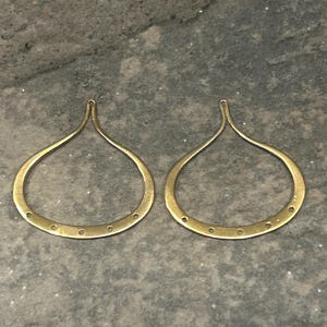 May include: Two gold-colored teardrop-shaped earring components. Each earring has a loop at the top and a series of small holes along the bottom curve. The earrings are displayed on a gray surface.