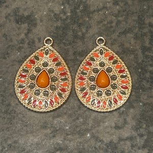 May include: Two gold-toned teardrop-shaped pendants. Each pendant features a central orange-colored stone surrounded by a pattern of red, orange, and green accents. The pendants have a decorative gold border and a small loop at the top.