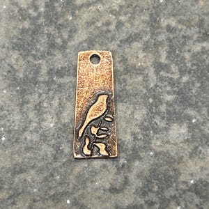 May include: A rectangular, bronze-colored pendant with a small hole at the top. The pendant features a detailed silhouette of a bird perched on a branch. The surface has a textured, aged appearance.