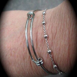 K&ouml;nnte beinhalten: Zwei silberne Armb&auml;nder an einem Handgelenk. Ein Armband ist ein einfaches Armreif mit einem Knotendetail. Das andere Armband ist eine Kette mit kleinen silbernen Perlen.