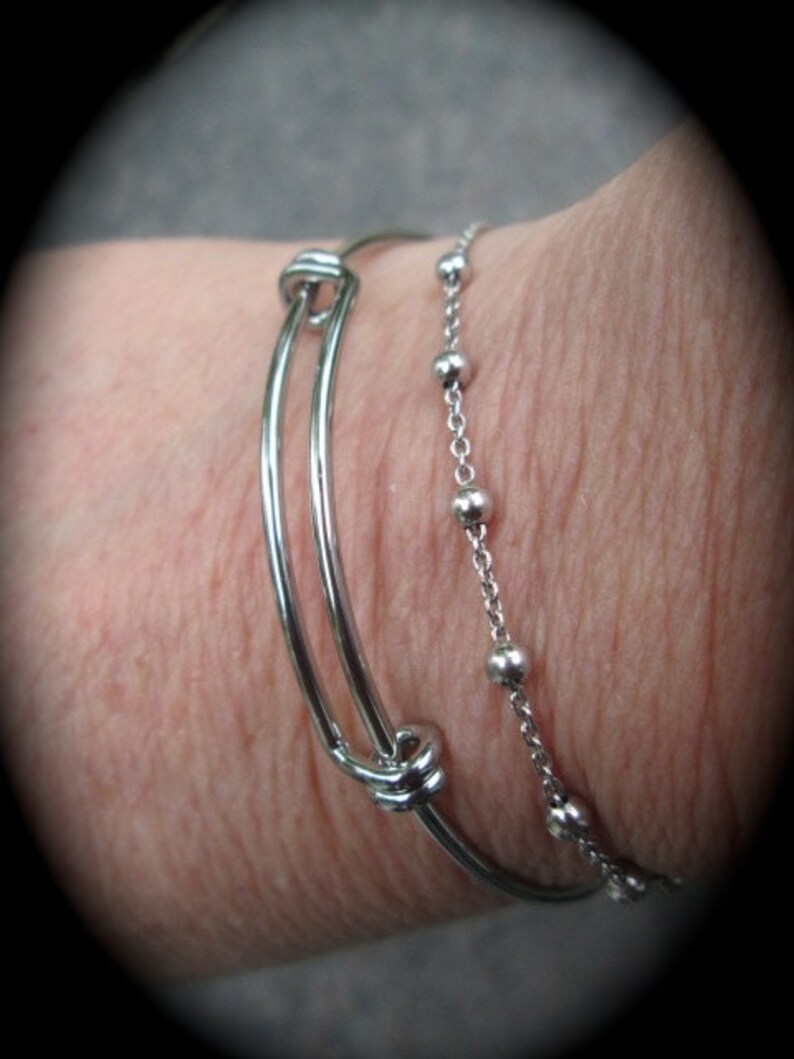 K&ouml;nnte beinhalten: Zwei silberne Armb&auml;nder an einem Handgelenk. Ein Armband ist ein einfaches Armreif mit einem Knotendetail. Das andere Armband ist eine Kette mit kleinen silbernen Perlen.