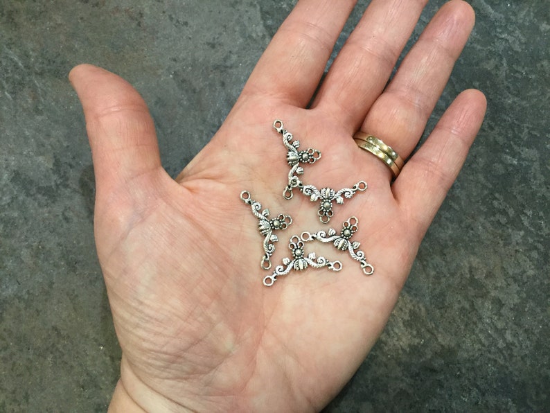 May include: Five silver-toned metal connector charms with a floral design. Each charm has three loops for attaching to jewelry or other crafts.