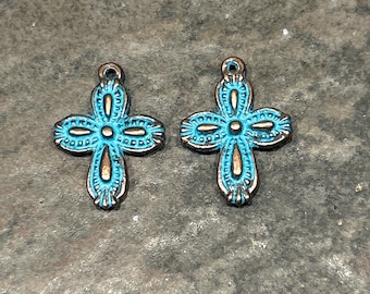 May include: Two turquoise cross-shaped pendants with a distressed finish. The crosses have a detailed design with a bronze-colored outline and a small loop at the top for hanging. The background is a textured gray surface.