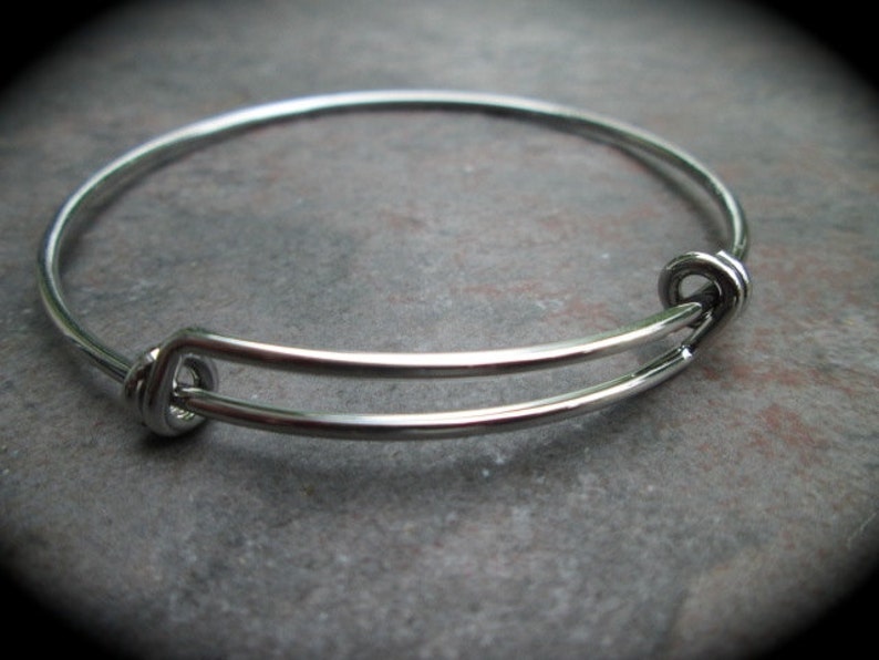 K&ouml;nnte beinhalten: Ein silbernes, verstellbares Armreif-Armband mit schlichtem Design. Das Armband ist aus Metall und hat eine glatte Oberfl&auml;che.