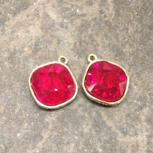 May include: Two gold-toned pendant charms with red faceted glass stones. The stones are square-shaped with rounded corners.