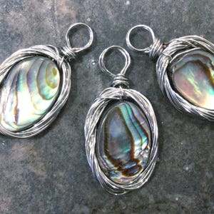 May include: Three oval pendants featuring iridescent abalone shell cabochons encased in silver-toned wire wrapping. Each pendant includes a circular wire bail at the top, showcasing swirling patterns of green, blue, and brown within the shell.