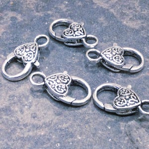May include: Five silver-toned metal lobster claw clasps arranged in a circular pattern. Each clasp features a heart-shaped base with an intricate, embossed filigree pattern on the front surface and a small circular loop at the top for attachment.