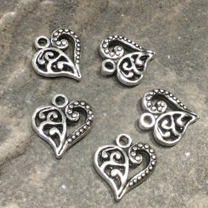 May include: Five silver-toned metal heart-shaped charms with intricate filigree designs. Each charm has a loop at the top for attaching to jewelry or other crafts.