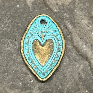 May include: A turquoise blue metal charm with a gold heart in the center. The heart is surrounded by a decorative border. The charm has a hole at the top for hanging.