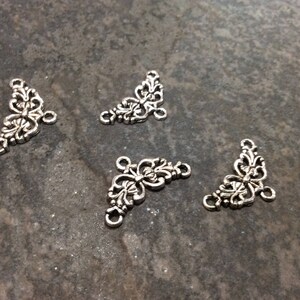 May include: Four silver metal corner connectors with a decorative floral design. Each connector has three holes for attaching to other jewelry pieces.